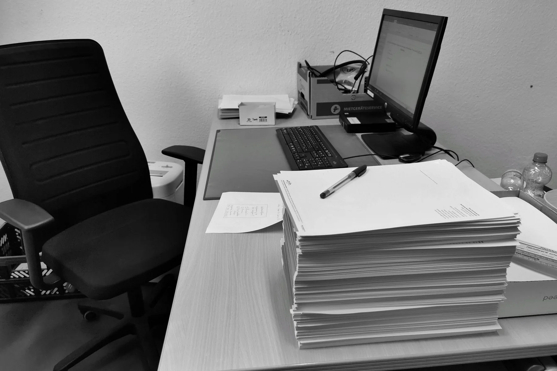 Black and white image of a cluttered office desk with a computer, chair, and paperwork.