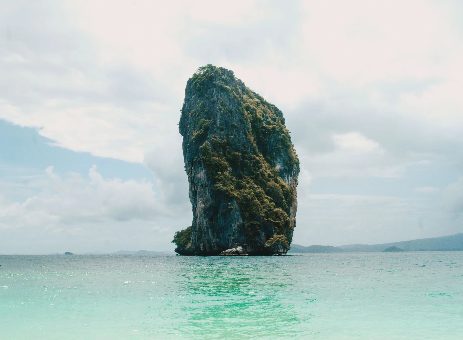 A striking limestone island emerges from the clear waters of Thailand, under a cloudy sky.